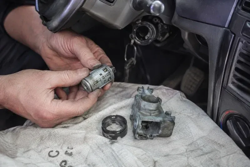 Hands holding a removed ignition cylinder beside a car steering column, with the housing and small parts on a cloth. — Ignition Lock Repair, Oakville